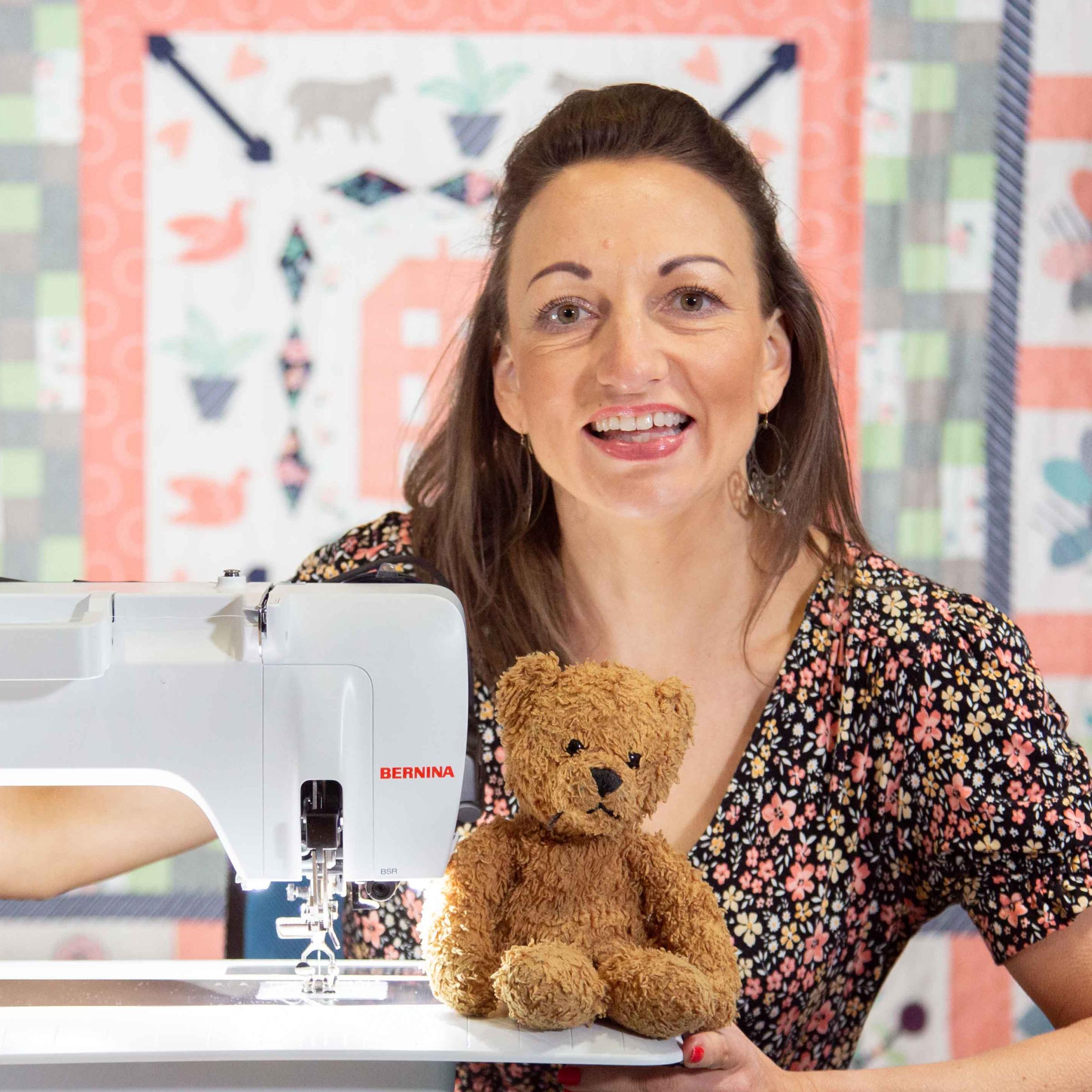 Quilter Sarah Ashford sits at a sewing machine smiling toward the camera, a colourful pieced patchwork quilt hangs as a backdrop and a teddy bear sits on the sewing machine.