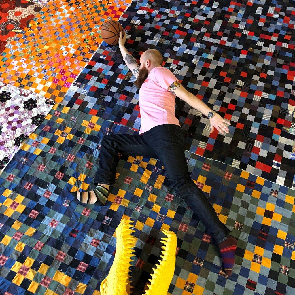 Quilter Chris English lies on a background of his own nine patch and hexagon patchwork quilts, in a pose as though he is leaping from the jaws of a yellow plastic crocodile at the bottom of the frame with a basketball poised to shoot in his right hand.