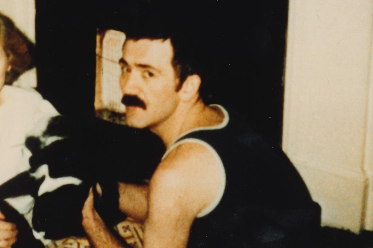 A young man with a moustache turns to look over his shoulder at the viewer, he is stroking a black and white cat.