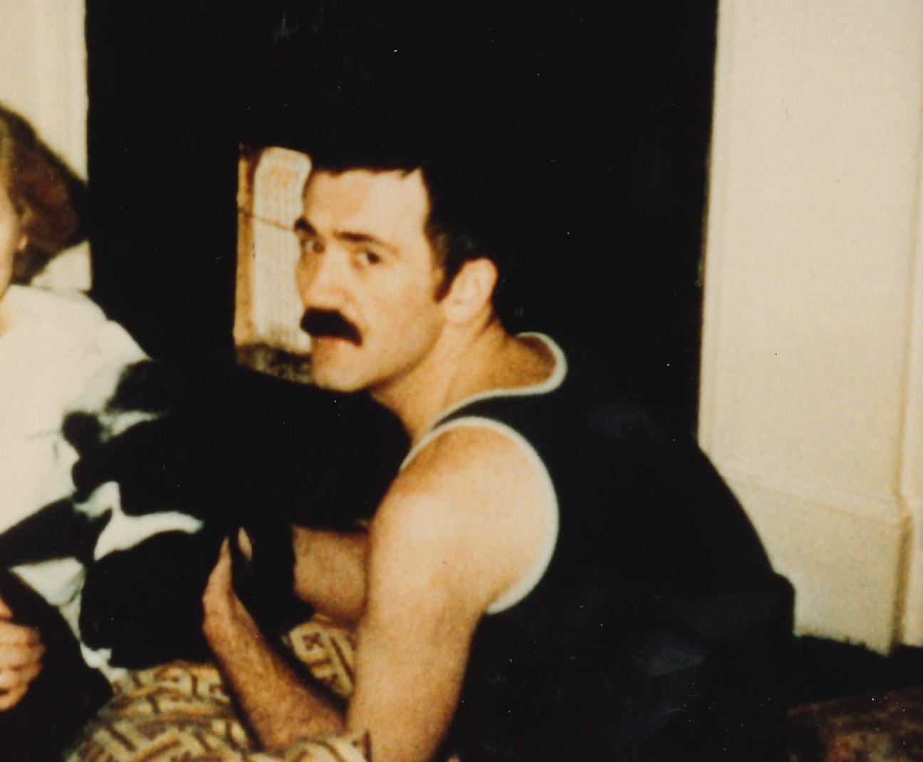 A young man with a moustache turns to look over his shoulder at the viewer, he is stroking a black and white cat.