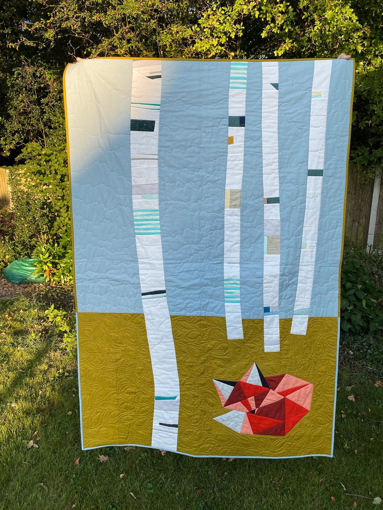 Image shows a modern quilt depicting a fox on a green ground in front of a grove of birch trees using solid colours and angular shapes.