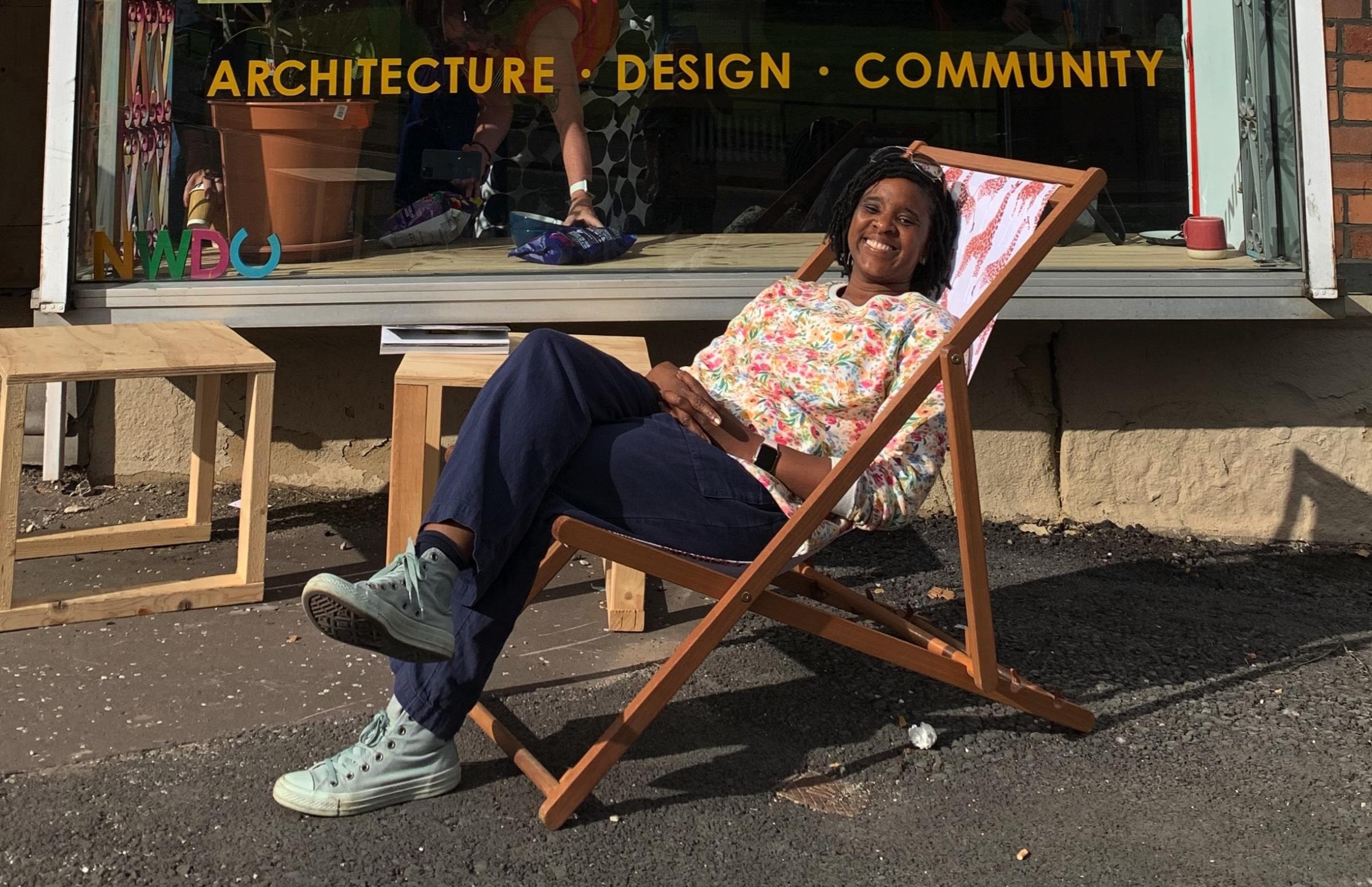 A smiling black woman with chin length black hair sits in a deckchair outside an architectural firm shop window.