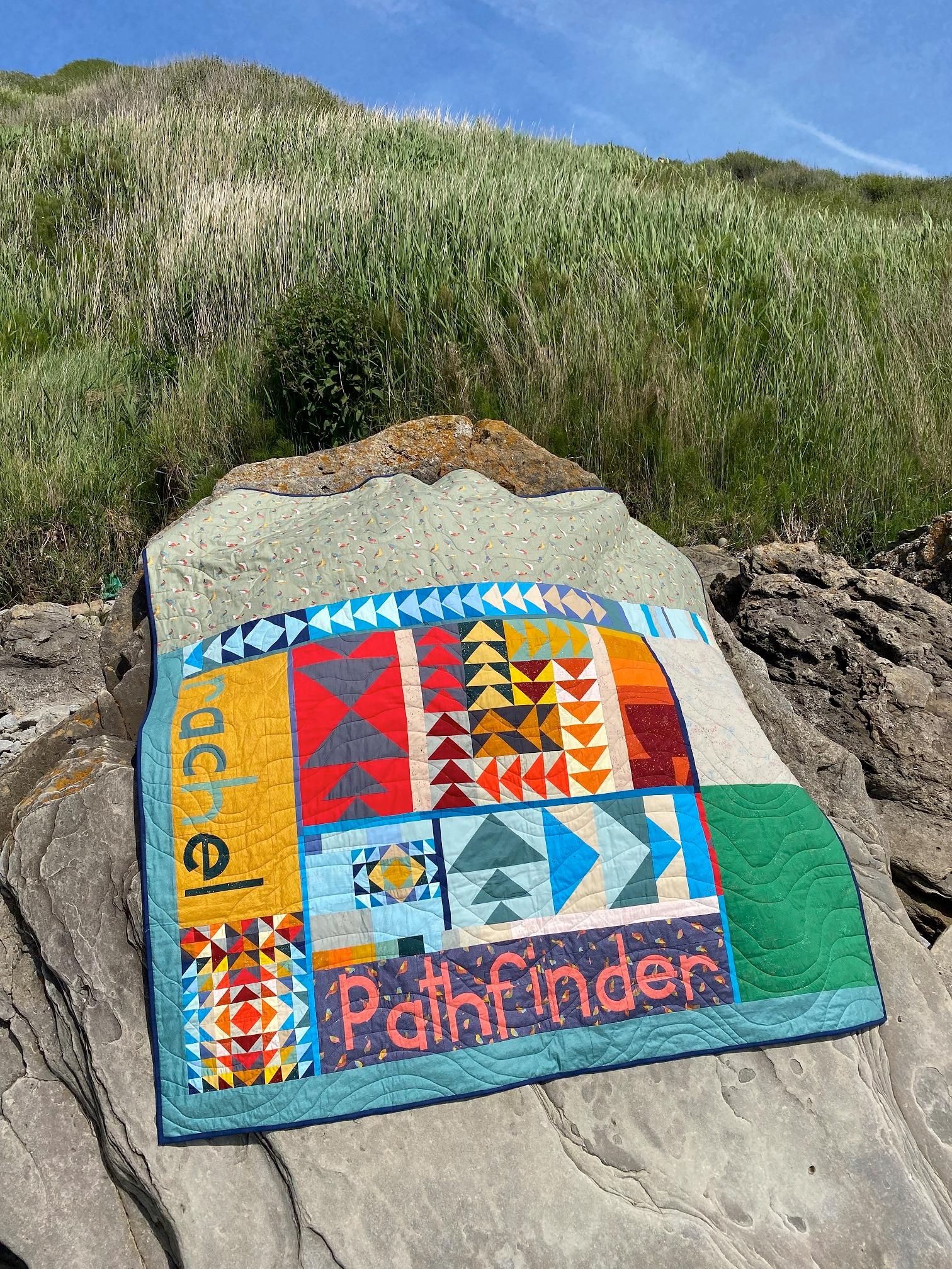 The reverse of a quilt made up of flying geese blocks in bright primary and earthy colours, and the names 'Rachel' and 'Pathfinder' lays on a rock on the beach, with sand dunes and blue sky behind.