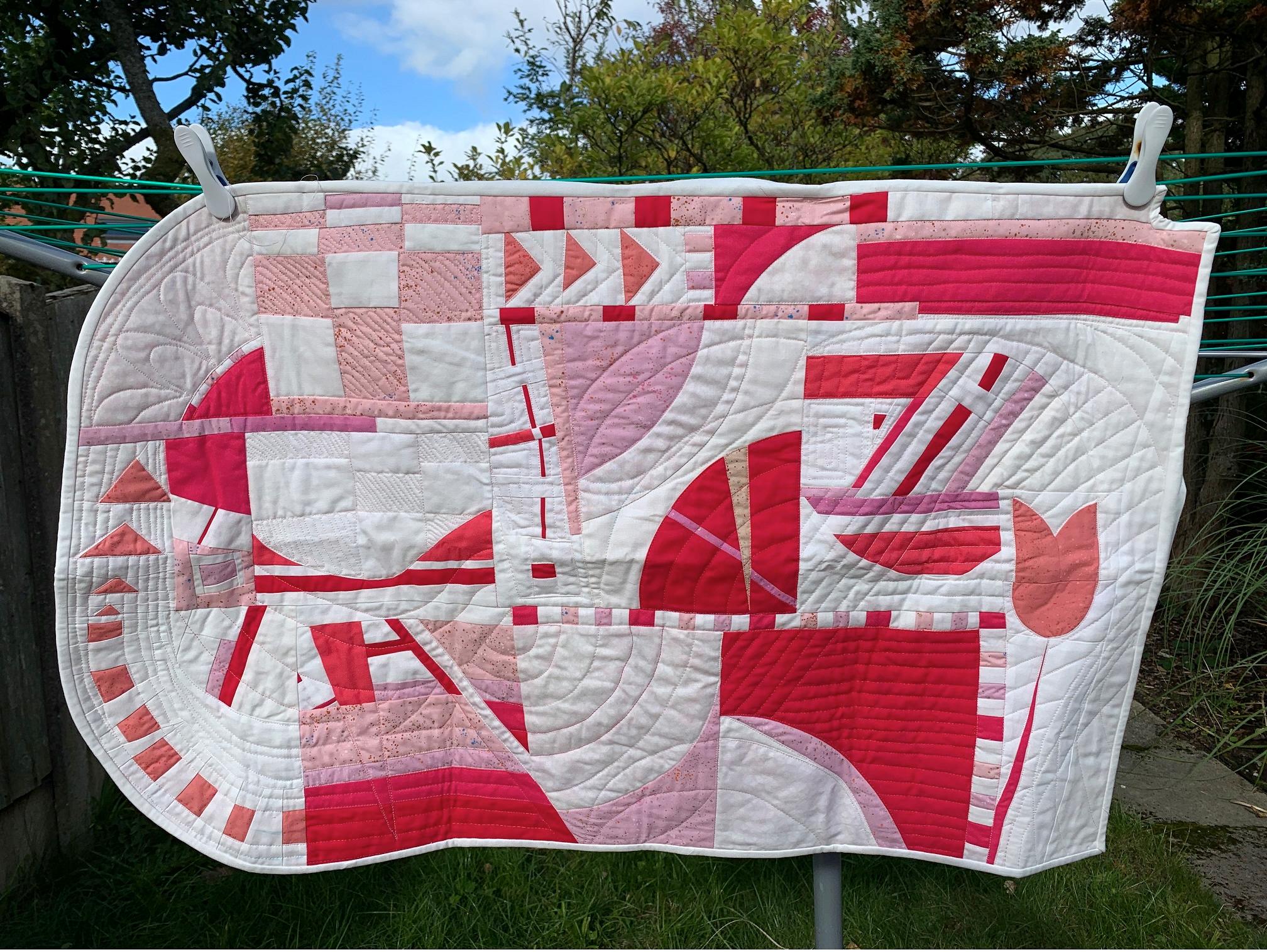 A modern abstract quilt in shades of pink and white hangs on a washing line, with trees and blue sky in the background.