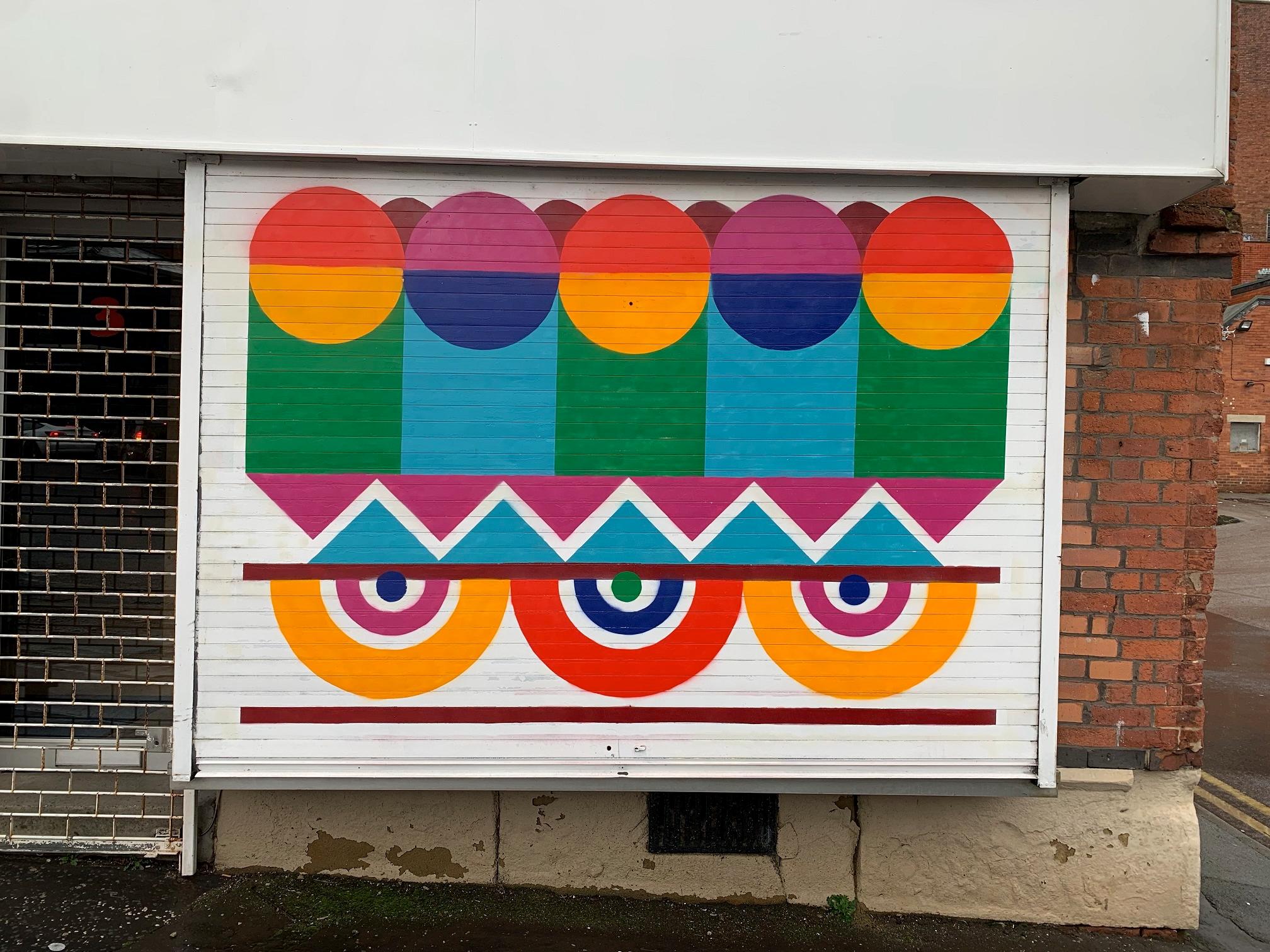 A white locked shop shutter bears an abstract design of circles, semicircles, triangles and lines in primary and secondary colours.
