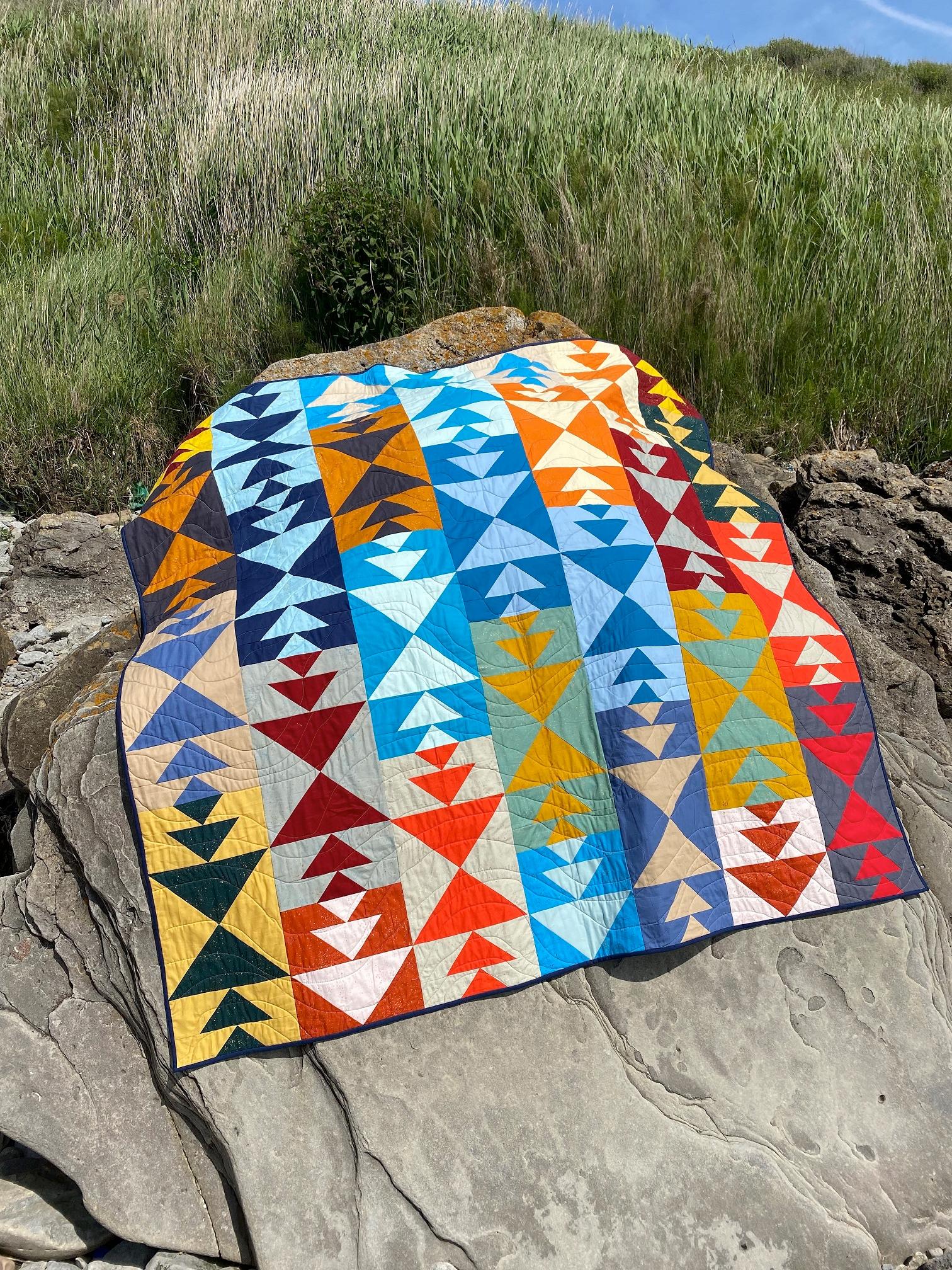 A quilt made up of flying geese blocks in bright primary and earthy colours lays on a rock on the beach, with sand dunes and blue sky behind.