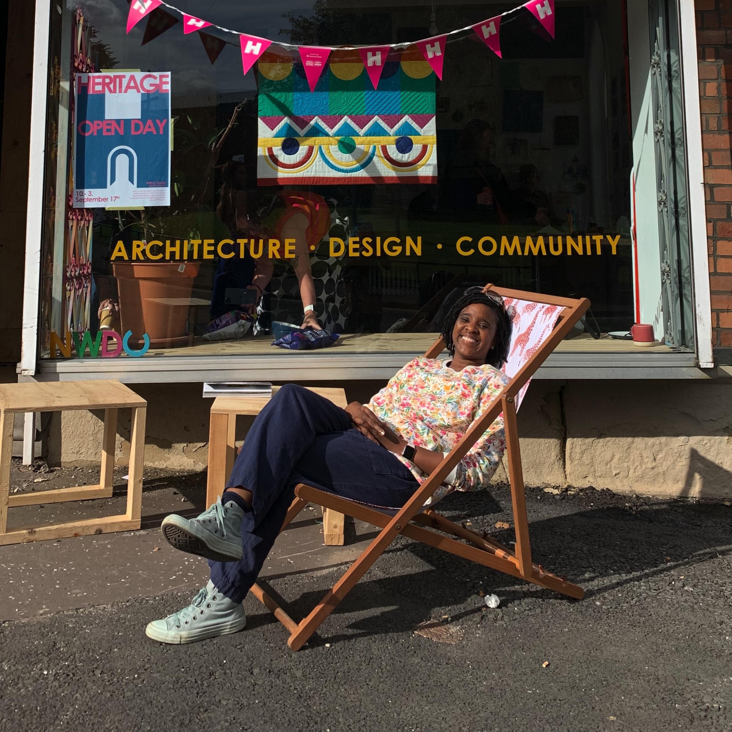 A smiling black woman with chin length black hair sits in a deckchair outside an architectural firm shop window. The shop window displays her abstract quilt in primary and secondary colours.