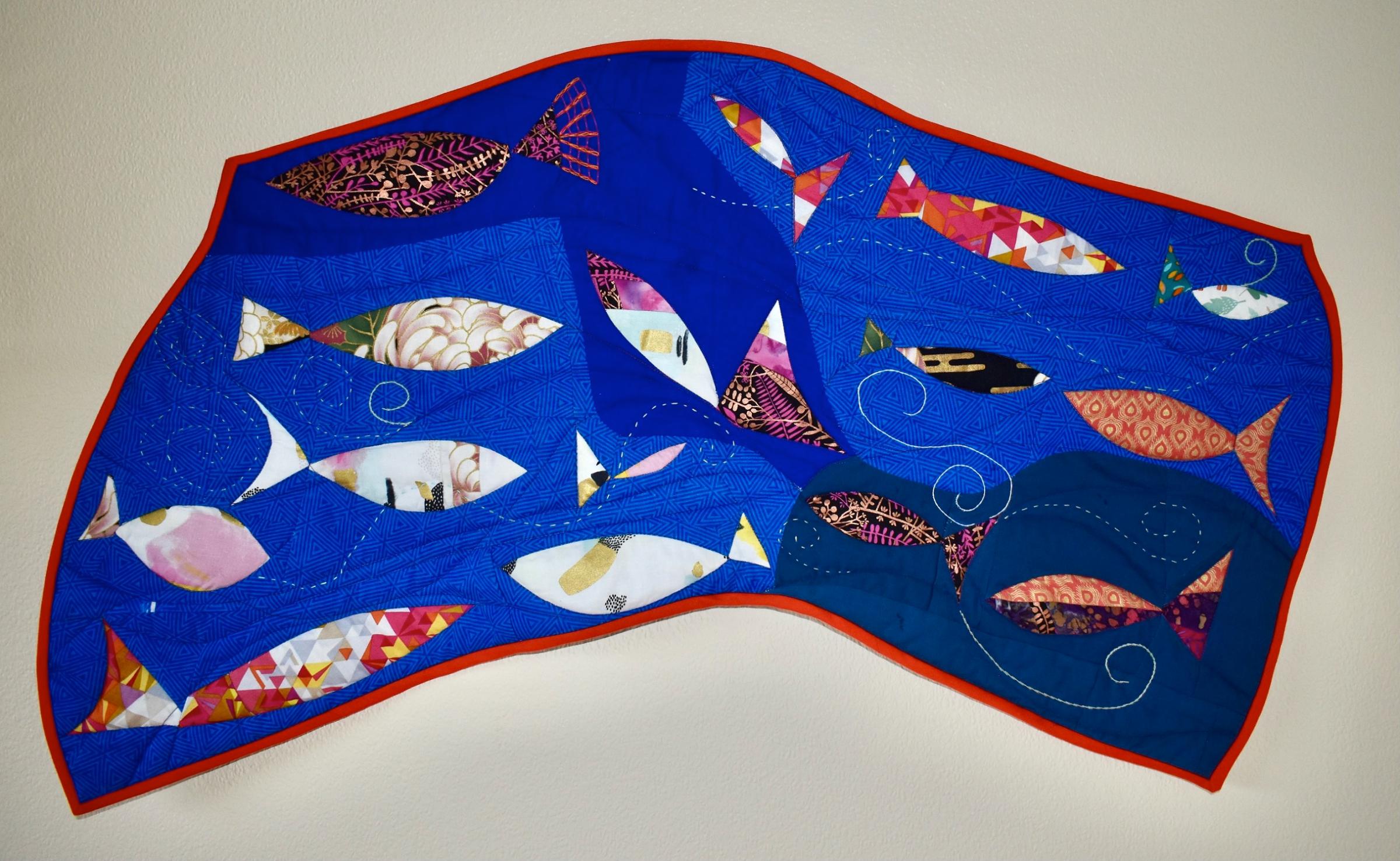 The image shows a modern quilt depicting stylised colourful fish on a blue background with red binding. The quilt is an irregular shape.