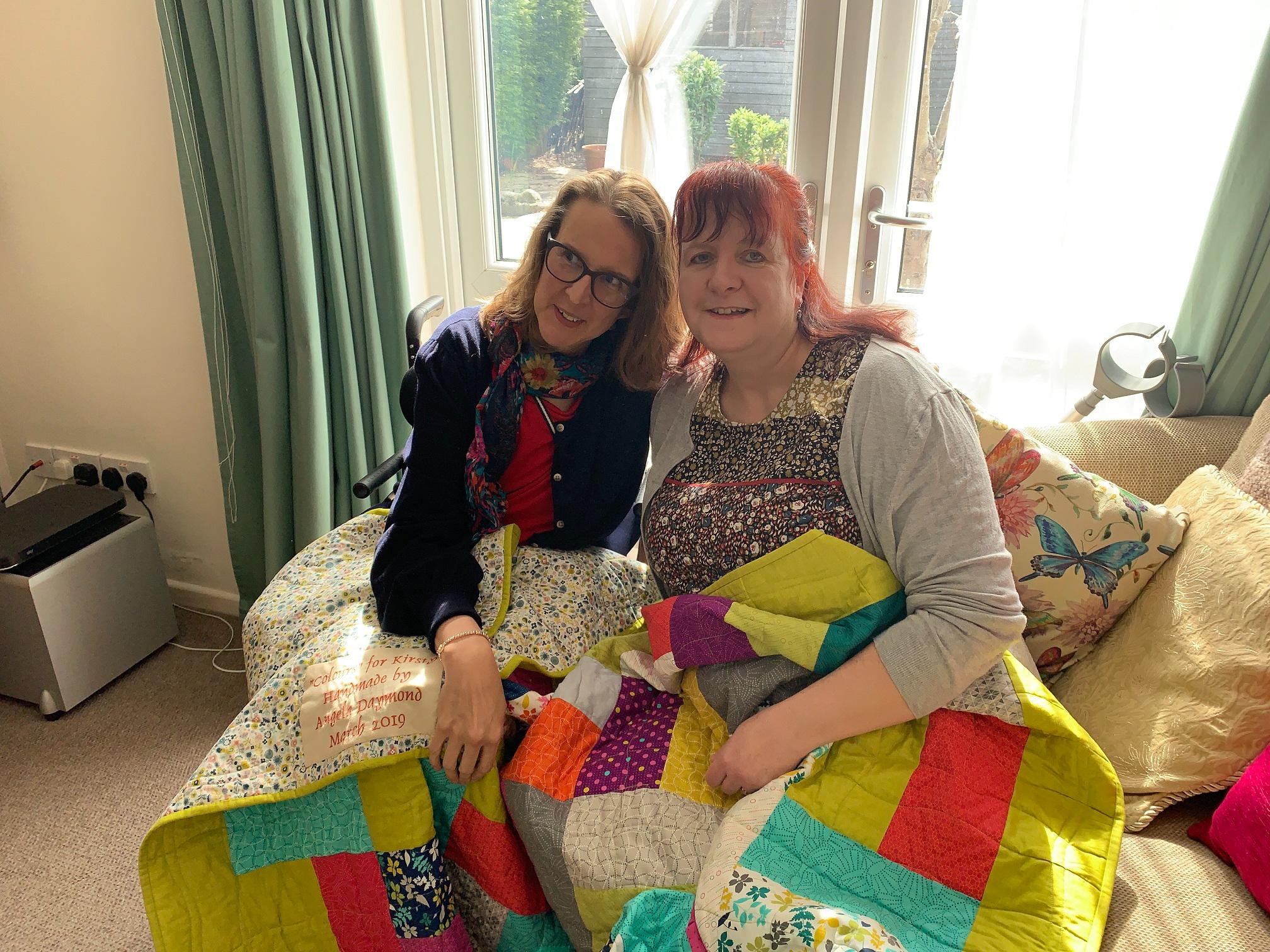 Two white women with long hair sit together underneath a colourful quilt. The woman on the left is in a wheelchair, the woman on the right is sat on a sofa.