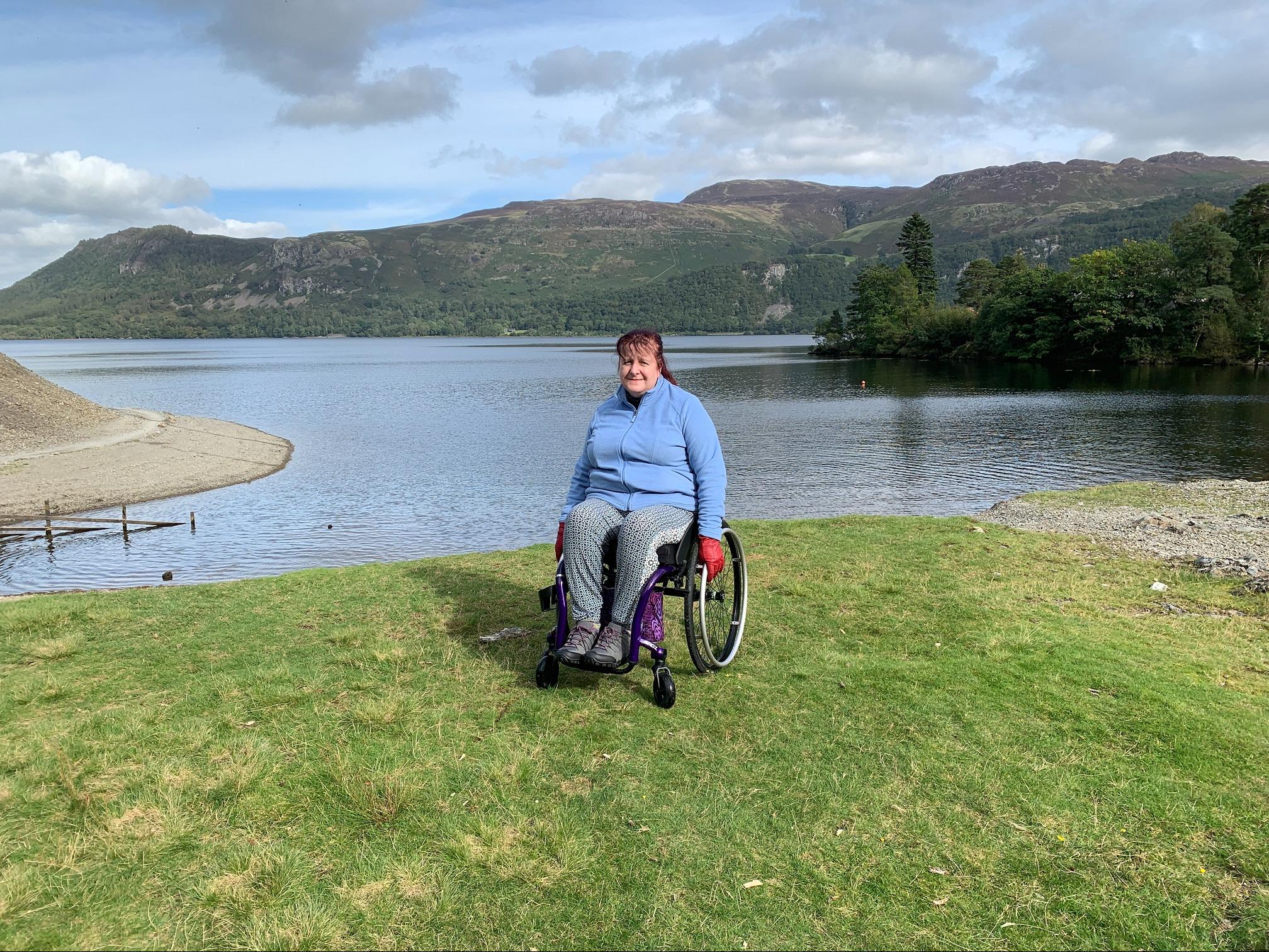 A white woman with mahogany red hair sits in a wheelchair on the shores of a lake, with hills in the distance behind her.