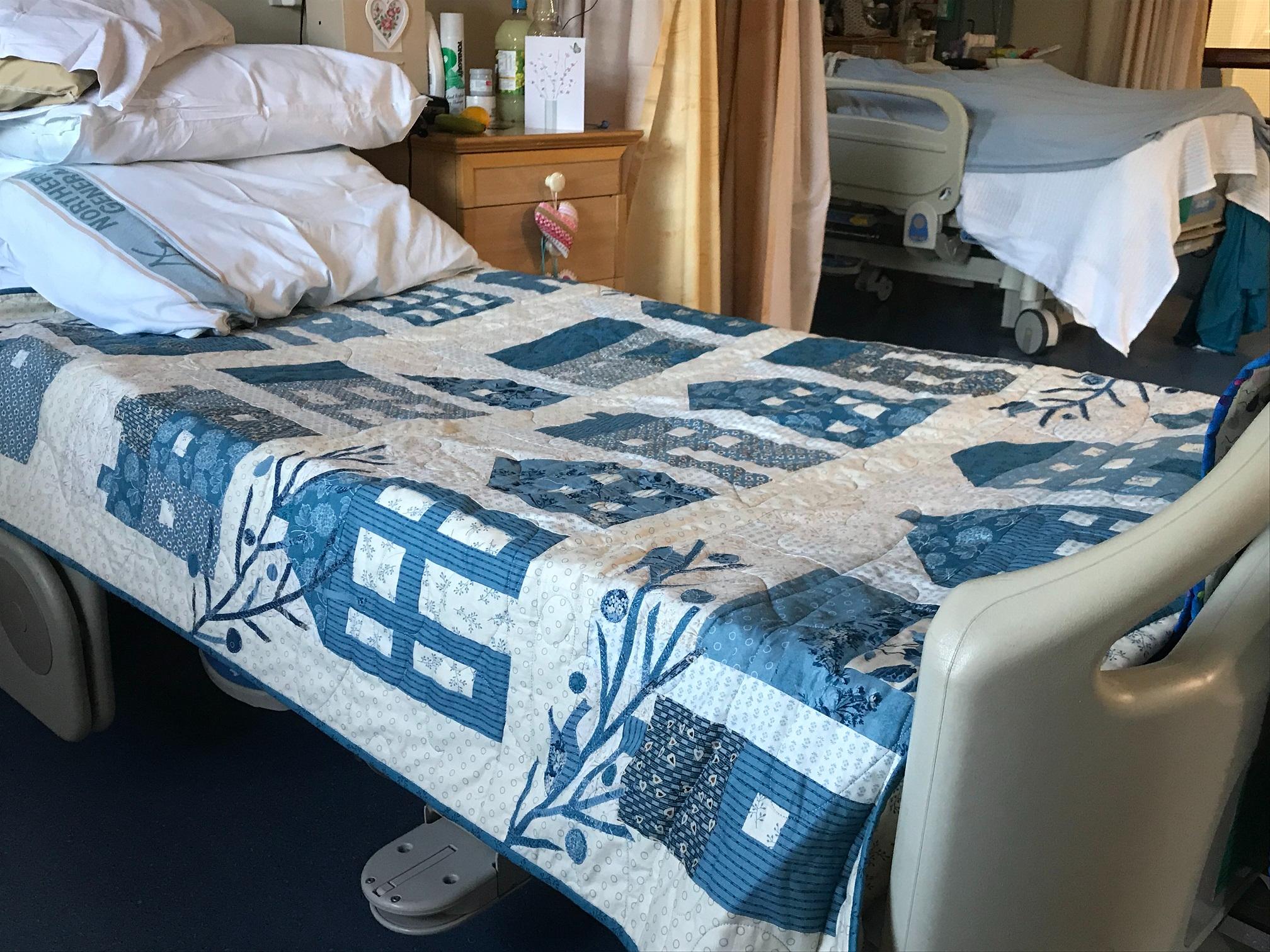 A blue and white quilt depicting different houses lies on a hospital bed. Another bed with a hospital blanket on it is in the background.