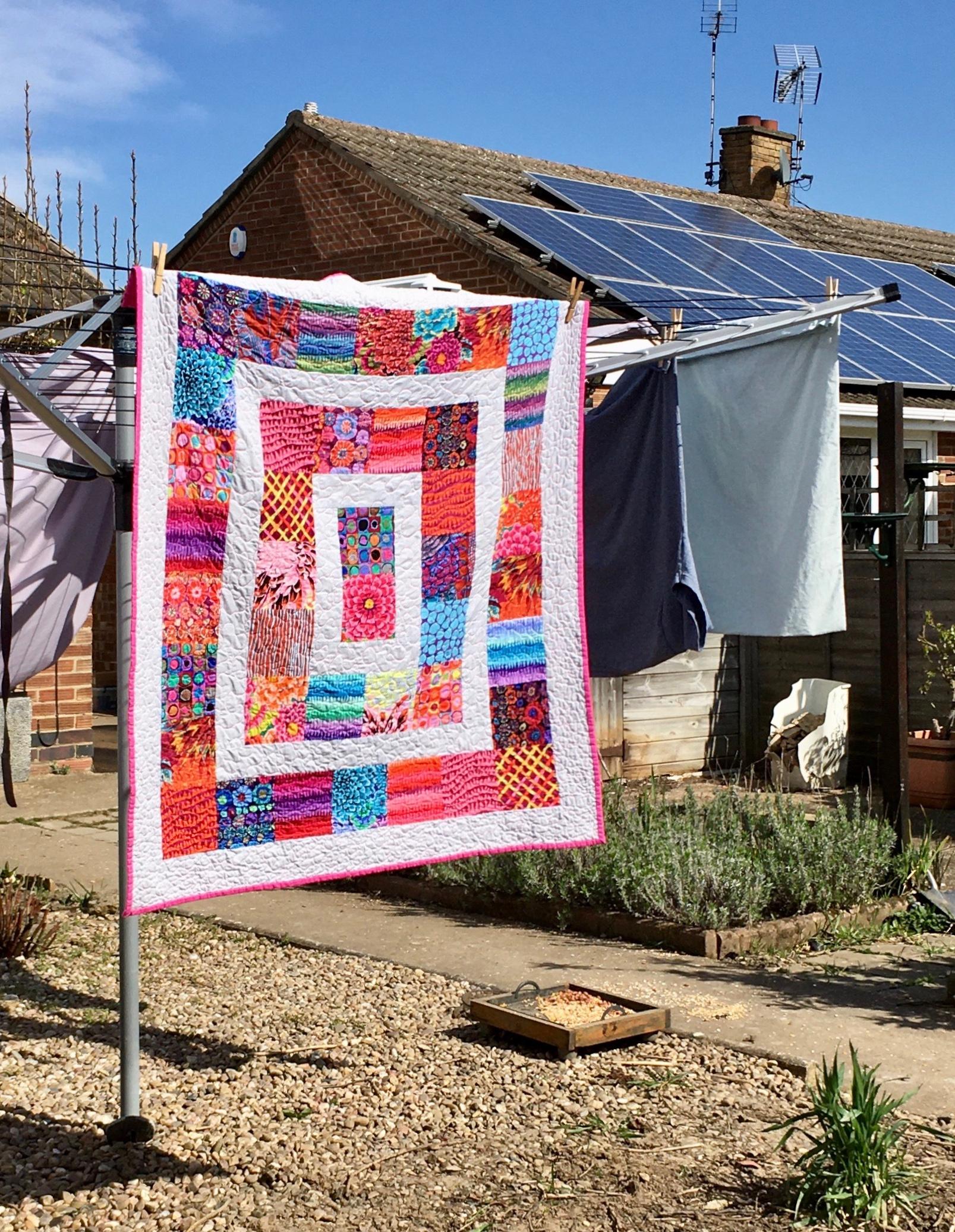 A frame quilt with alternating frames of white and brightly patterned fabrics in hues of red, orange, pink, blue, purple and green hangs on a washing line in a garden. A bungalow is in the background.