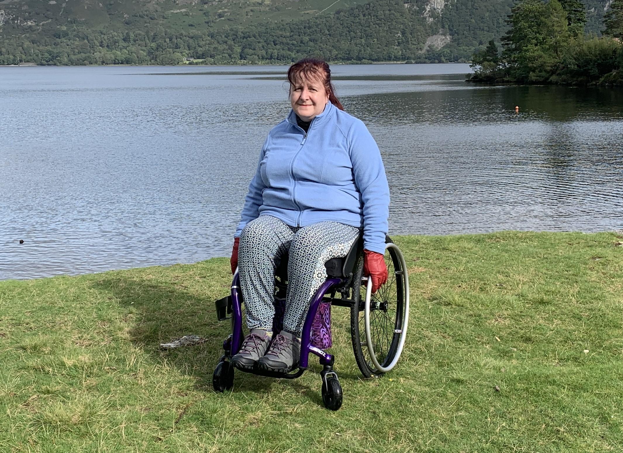 A white woman with mahogany red hair sits in a wheelchair on the shores of a lake.