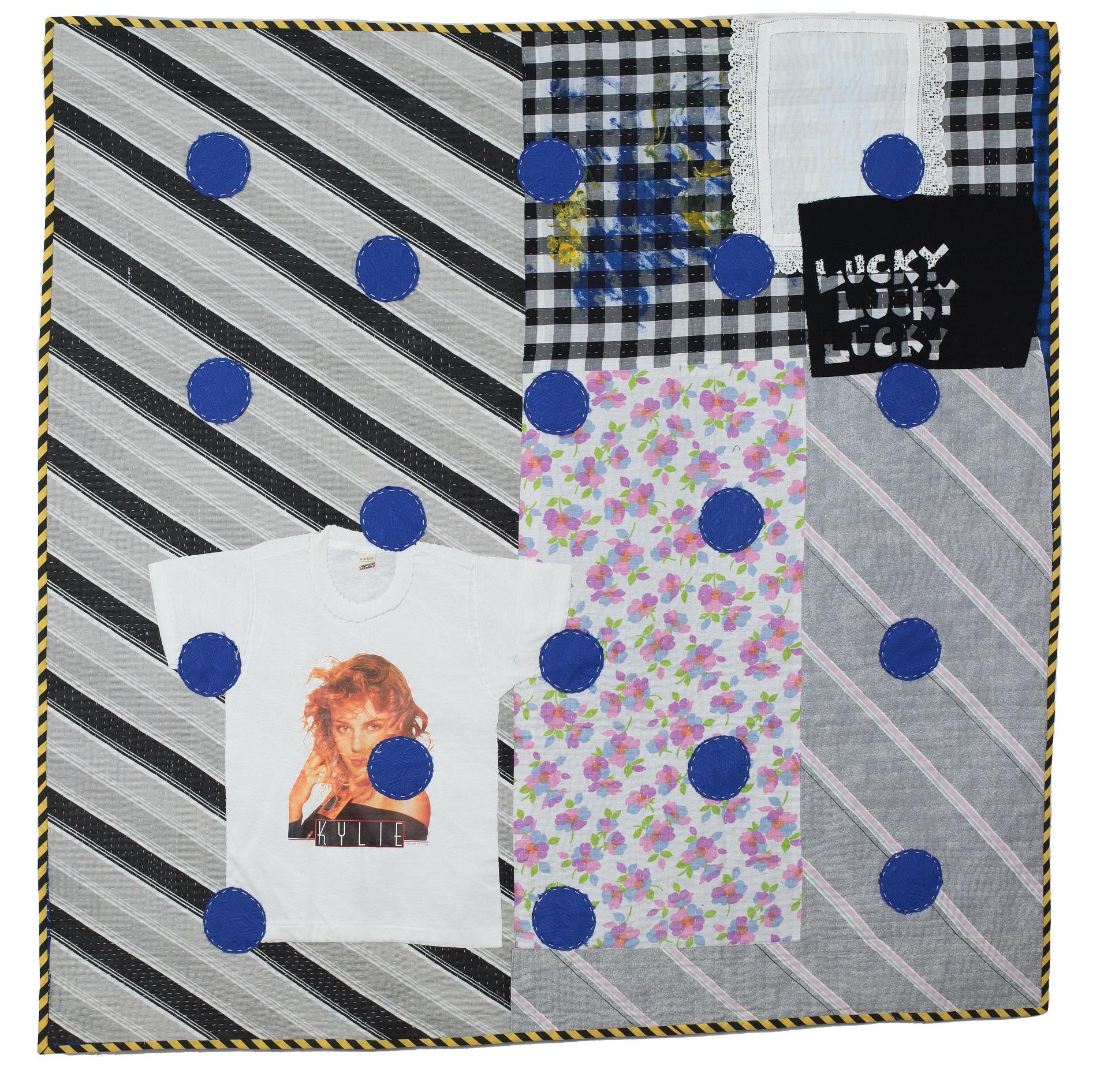 A modern quilt featuring greys and blacks and a pop of pink florals, a vintage Kylie Minogue t-shirt is appliqued onto the quilt, along with a vintage lace doily and the words 'Lucky, Lucky, Lucky'.