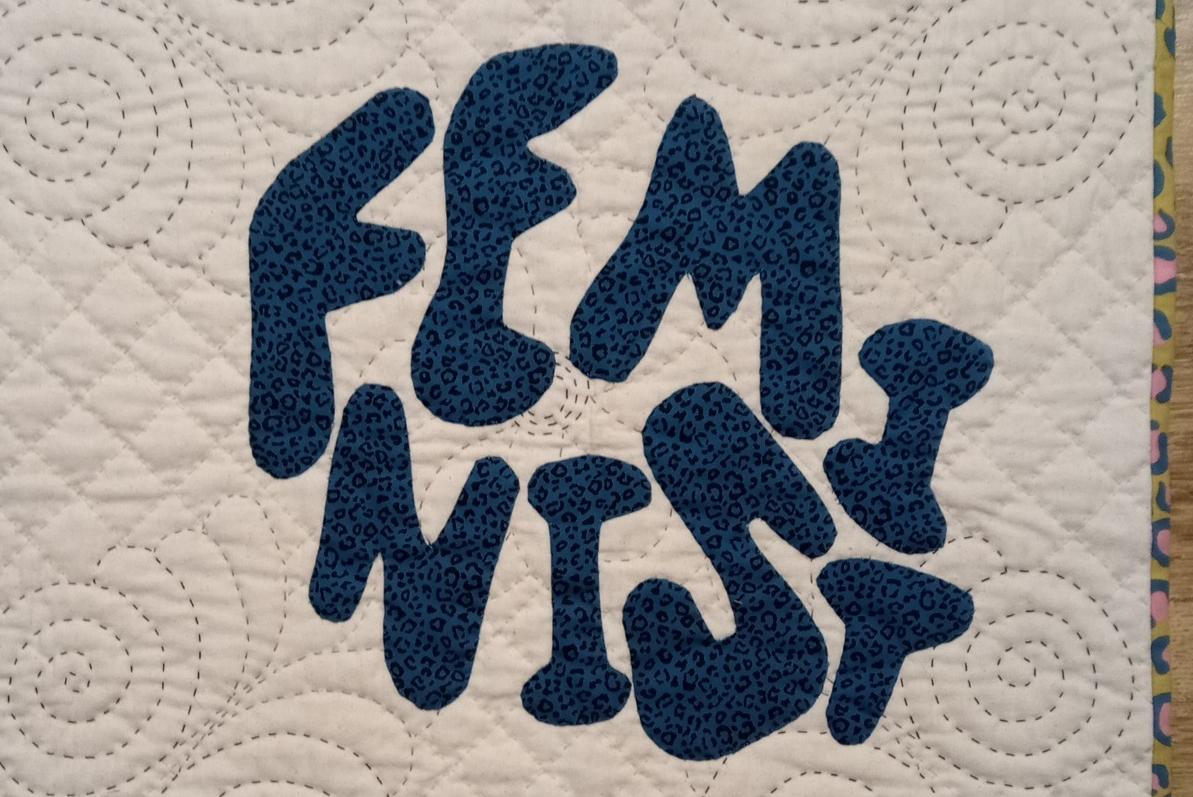 Image shows a quilt with white wholecloth background quilted with spirals and diamonds, and the word 'feminist' appliqued on in blue leopard print fabric.