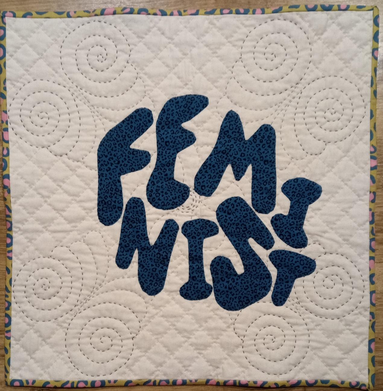 Image shows a quilt with white wholecloth background quilted with spirals and diamonds, and the word 'feminist' appliqued on in blue leopard print fabric and bound with a yellow leopard print.