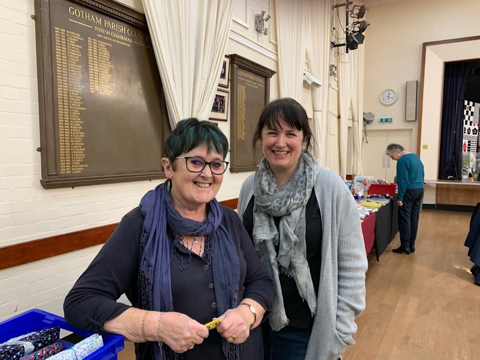 Two white women stand in a parish hall smiling at the camera, the woman in the foreground wears glasses and has short dark hair streaked with teal, and the woman in the background has long dark hair tied back.