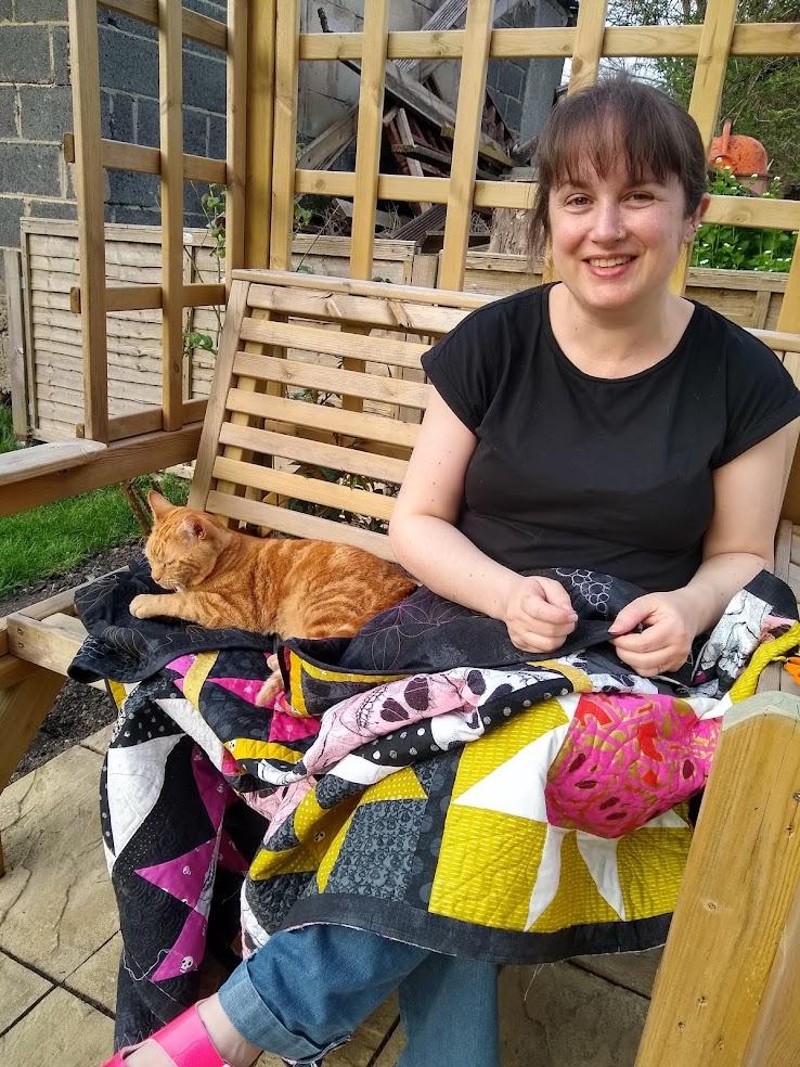 A white woman with brown hair sits on a swing bench in a garden, sewing a quilt on her lap, with a tabby cat sitting beside her.