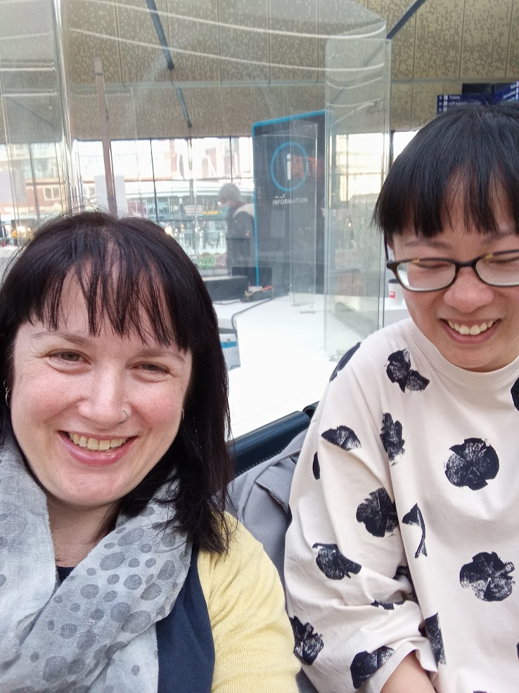 A white woman with dark hair and a nose ring, and a woman of east asian appearance wearing glasses, smile at the camera.