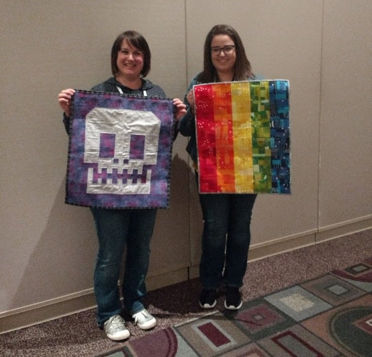 Image shows two white women smiling at the camera while holding up small quilts they have swapped with each other.