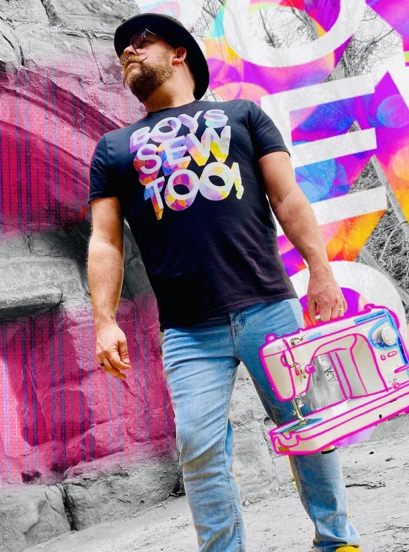 Image shows a white man with a beard, glasses and hat wearing a Boys Sew Too t-shirt in front of a wall of neon colour, he holds a sewing machine in his hand.