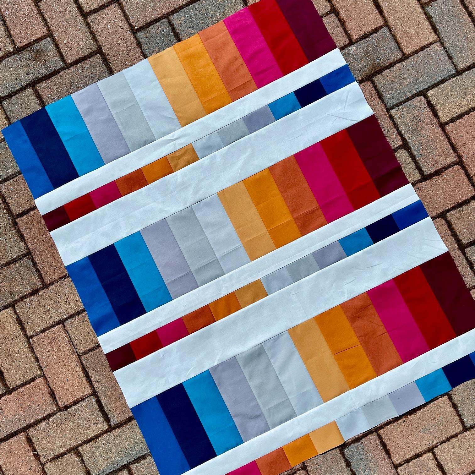 Image shows a pieced quilt top of graded colour strips, beginning with reds through oranges, yellows, grey and blues