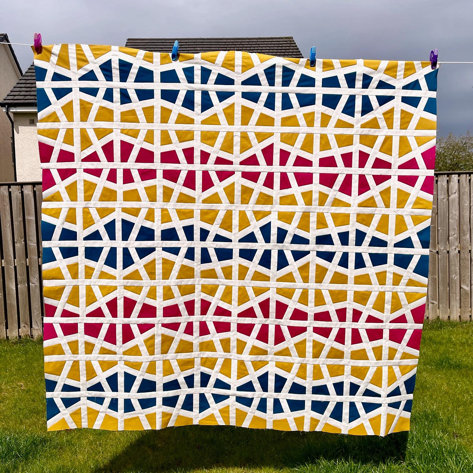 A red, yellow, white and blue geometric quilt top hangs on a washing line in a garden