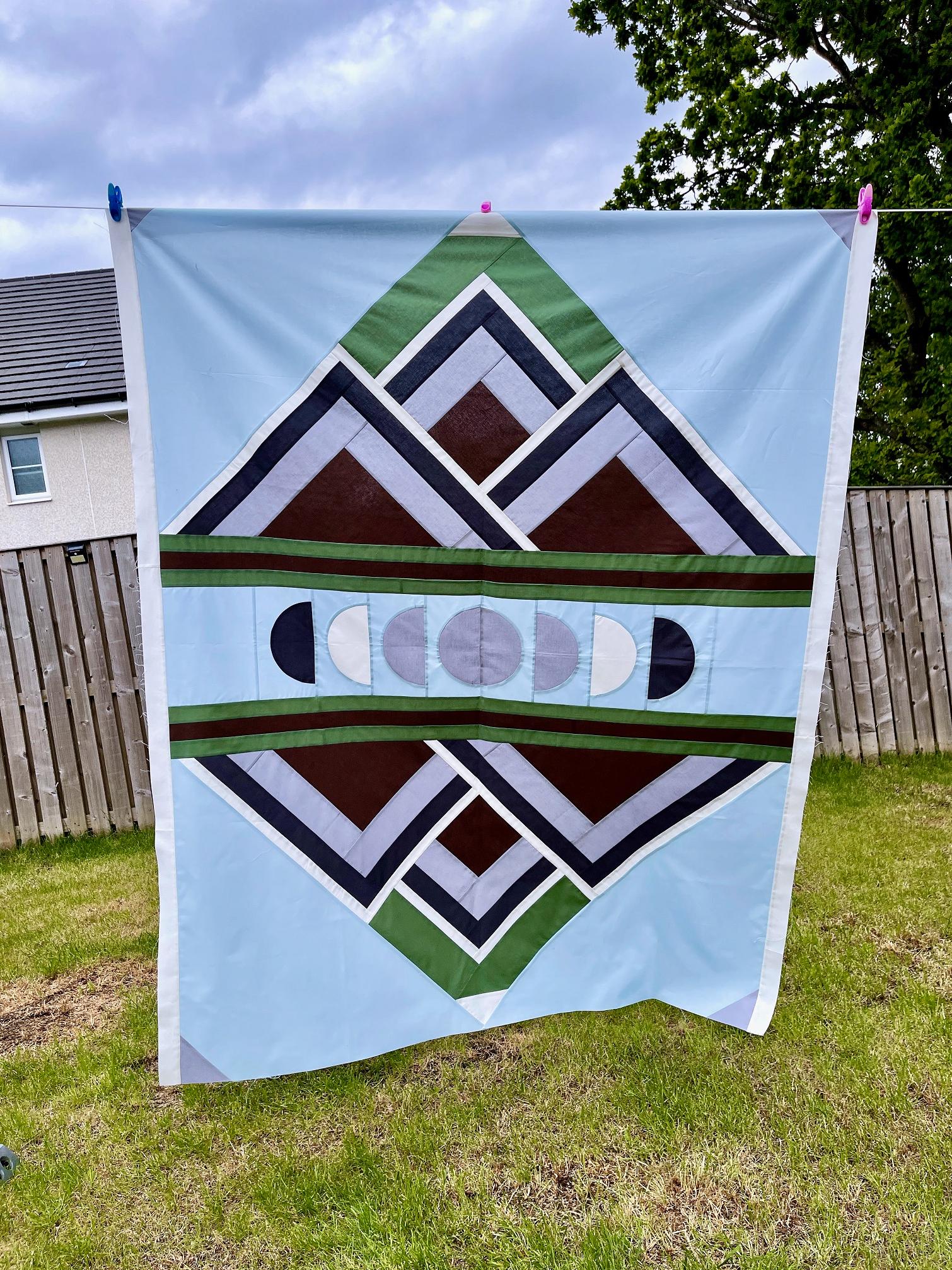 Image shows a mirror image quilt top featuring semi circles and triangular piecing in black, brown, grey, white and green on a pale blue background