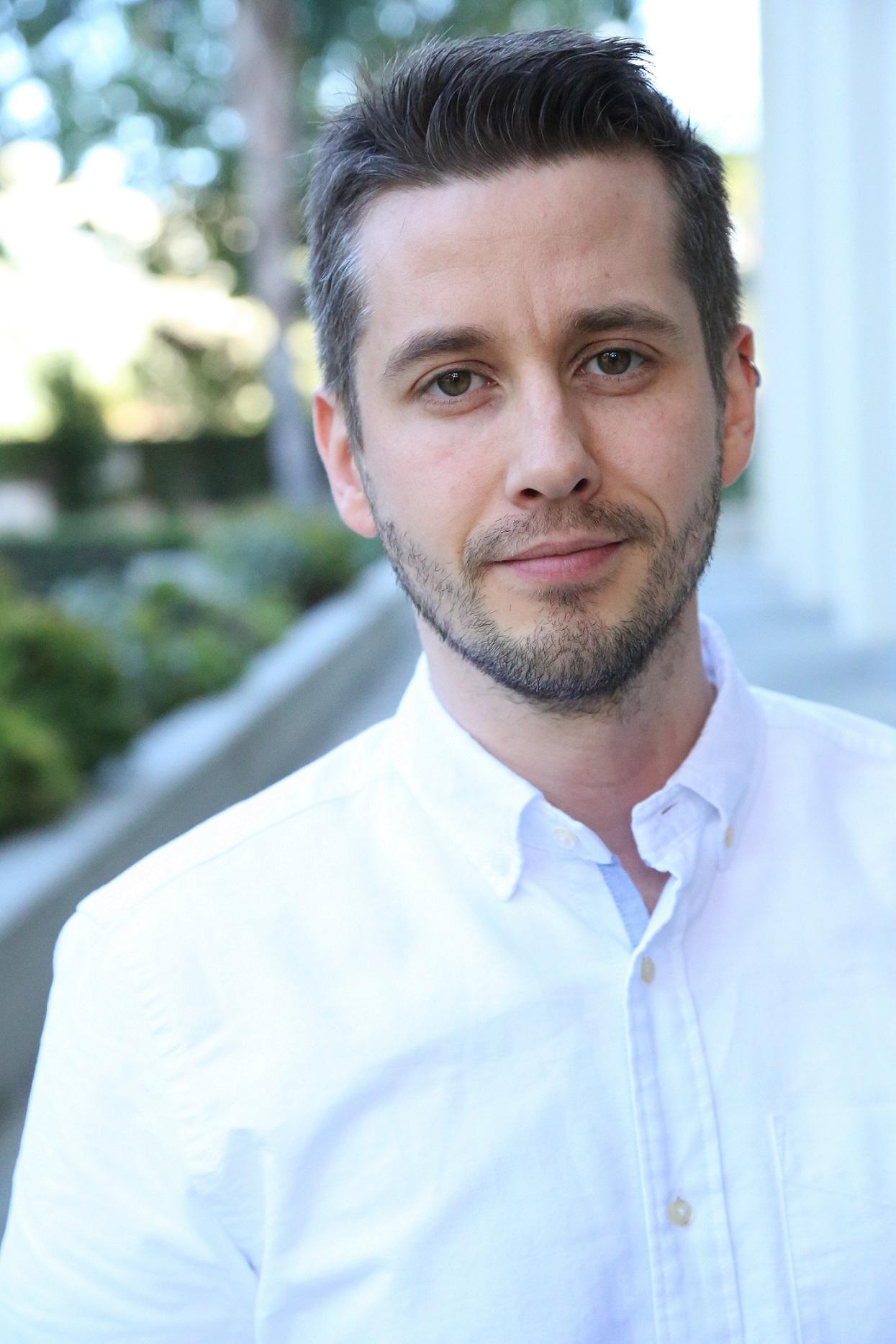 Image shows a white man with dark hair, light beard and glasses, wearing a white shirt