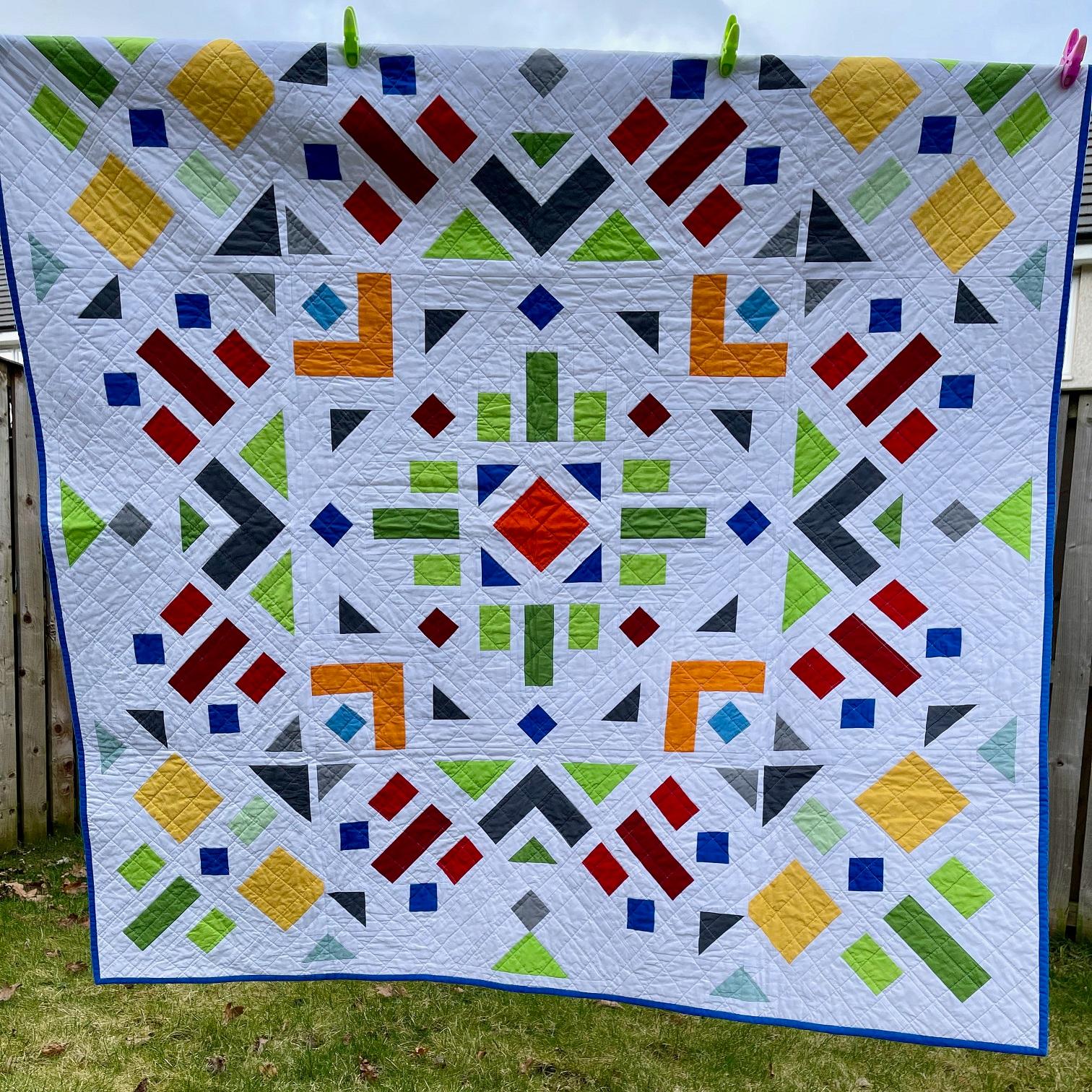 Image shows a quilt with a white background featuring geometric symmetrical shapes hanging on a washing line