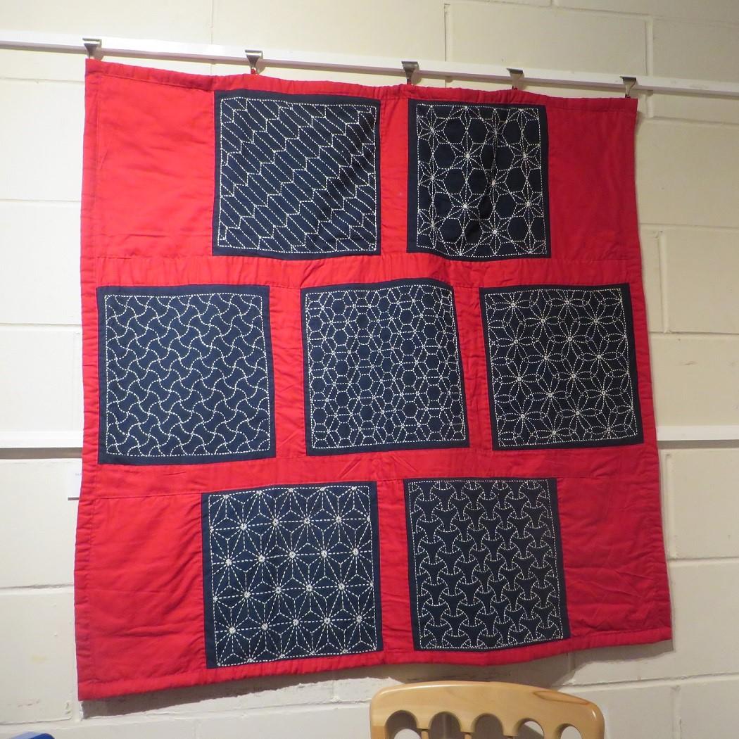 A square quilt with a red background features seven sashiko stitched squares, using white thread on black fabric.