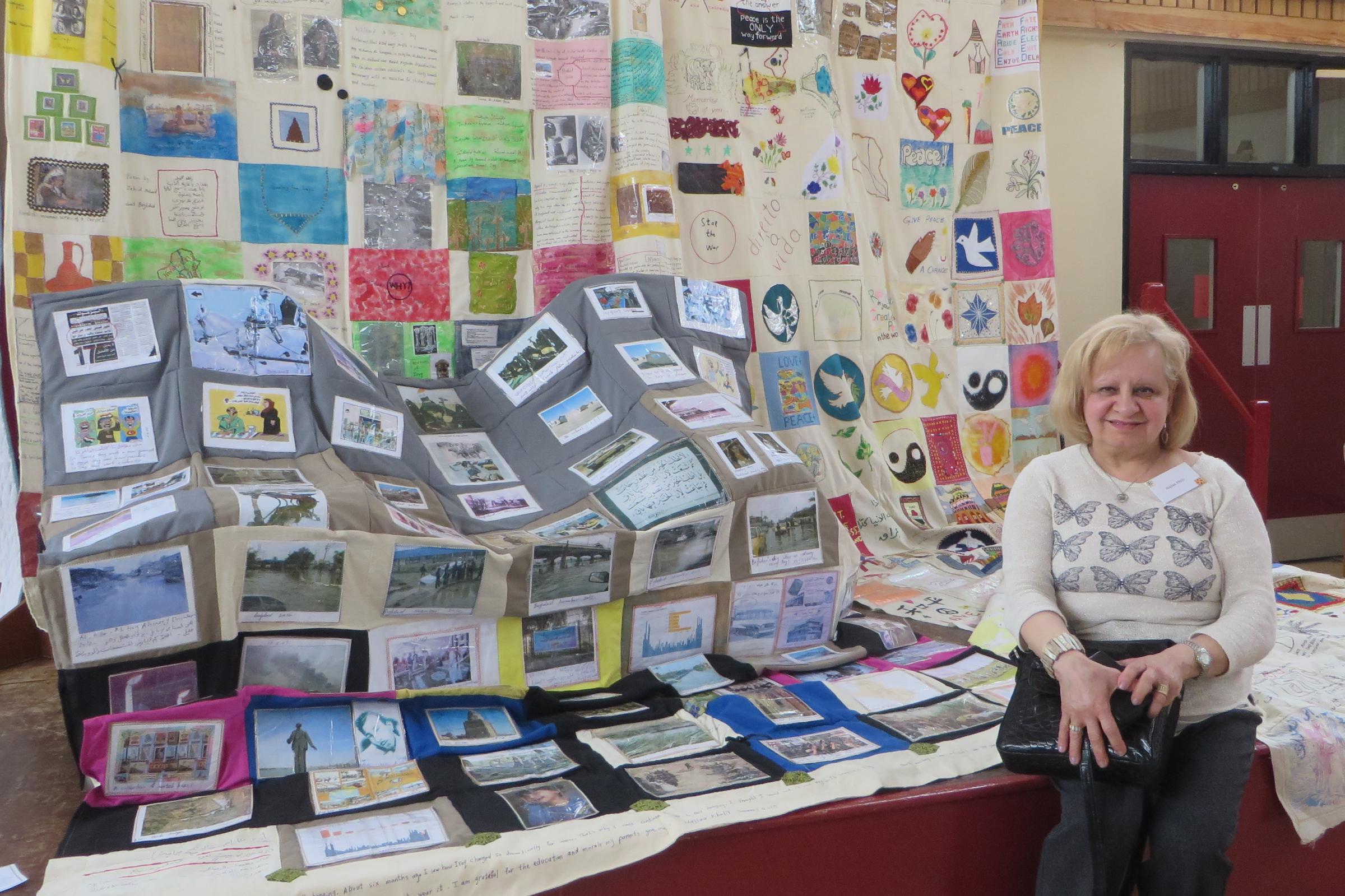 A smiling woman with fair hair sits in front of a display of her 'Piece for Peace' quilt project, made up of squares of cream canvas embroidered and appliqued with text and images