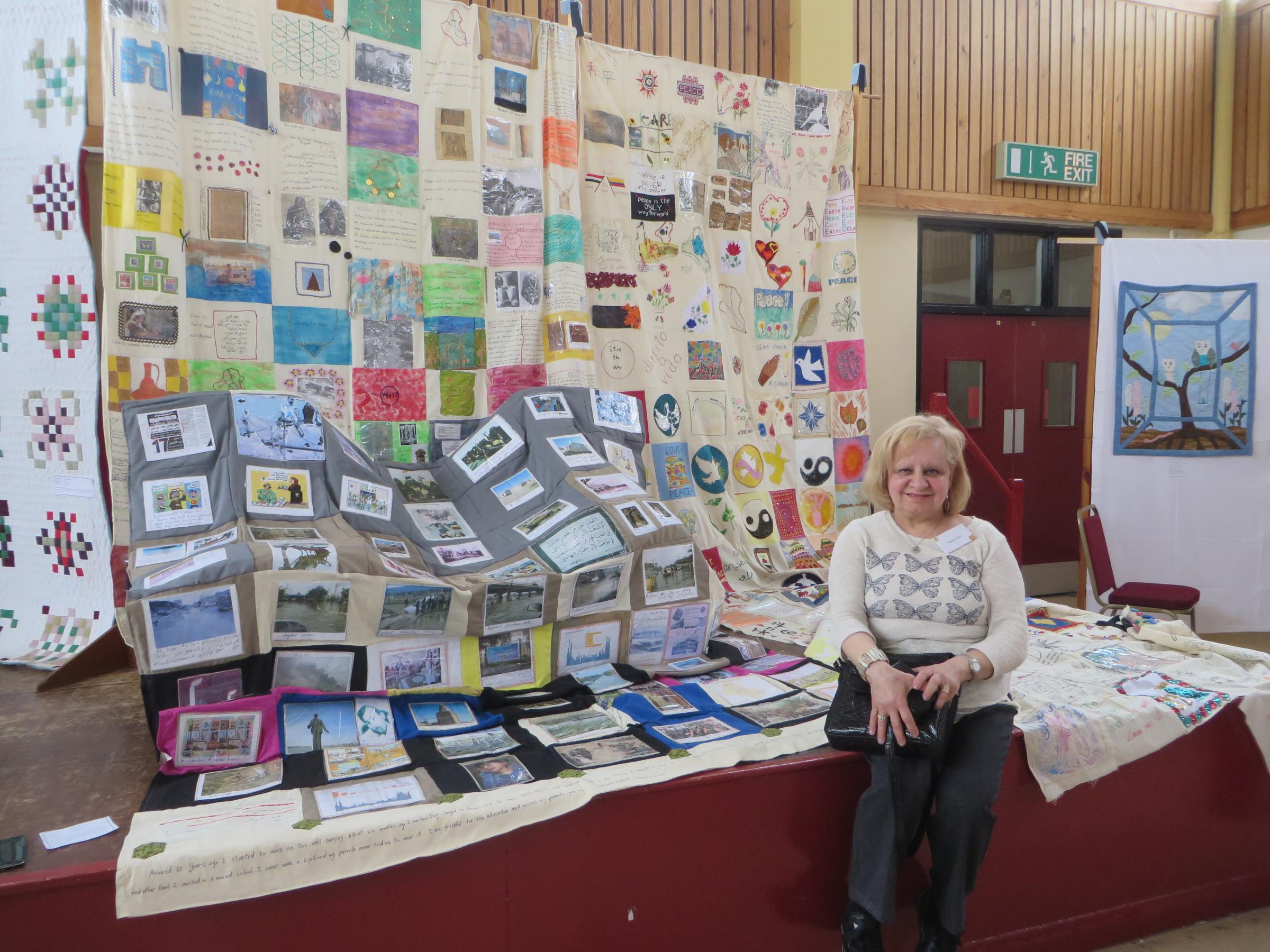 A smiling woman with fair hair sits in front of a display of her 'Piece for Peace' quilt project, made up of squares of cream canvas embroidered and appliqued with text and images