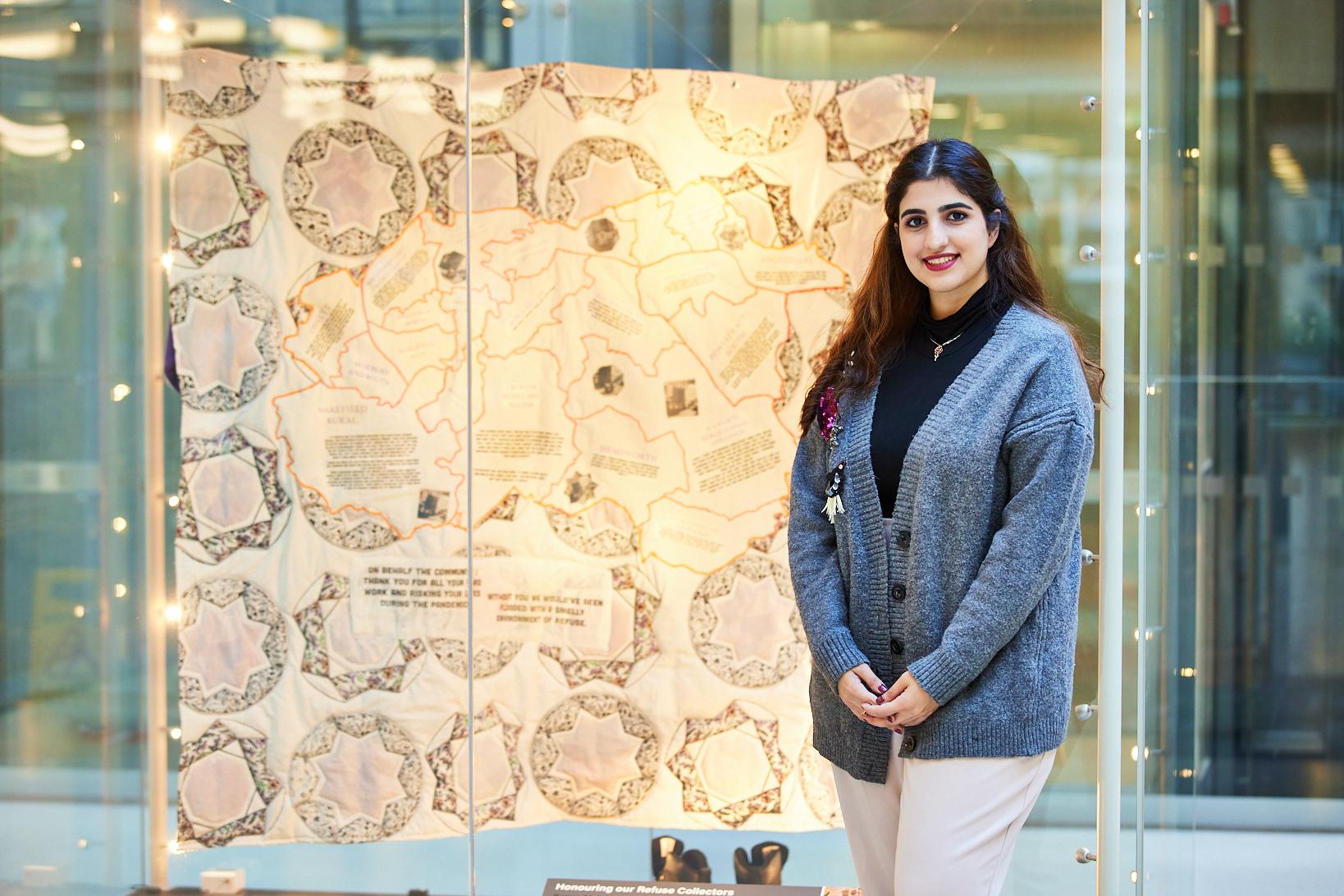 A woman with dark hair wearing a blue cardigan smiles at the camera whilst standing in front of a quilt bearing a map.