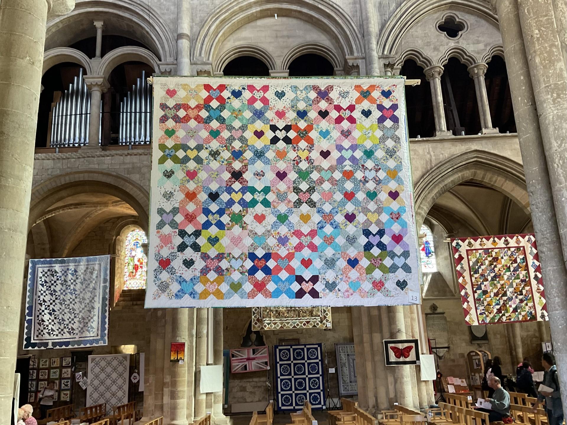 Image shows several exhibition quilts hanging suspended in the high gothic arches of a cathedral nave