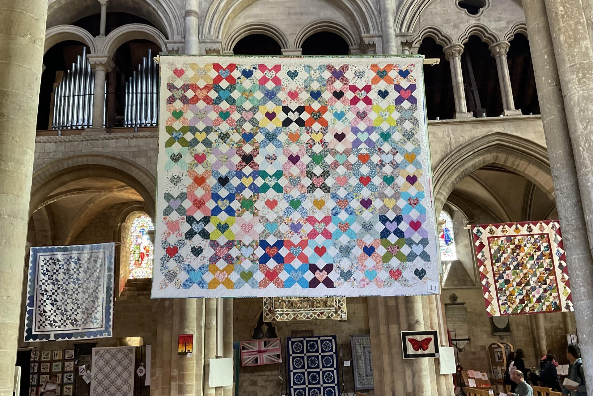 Image shows several exhibition quilts hanging suspended in the high gothic arches of a cathedral nave