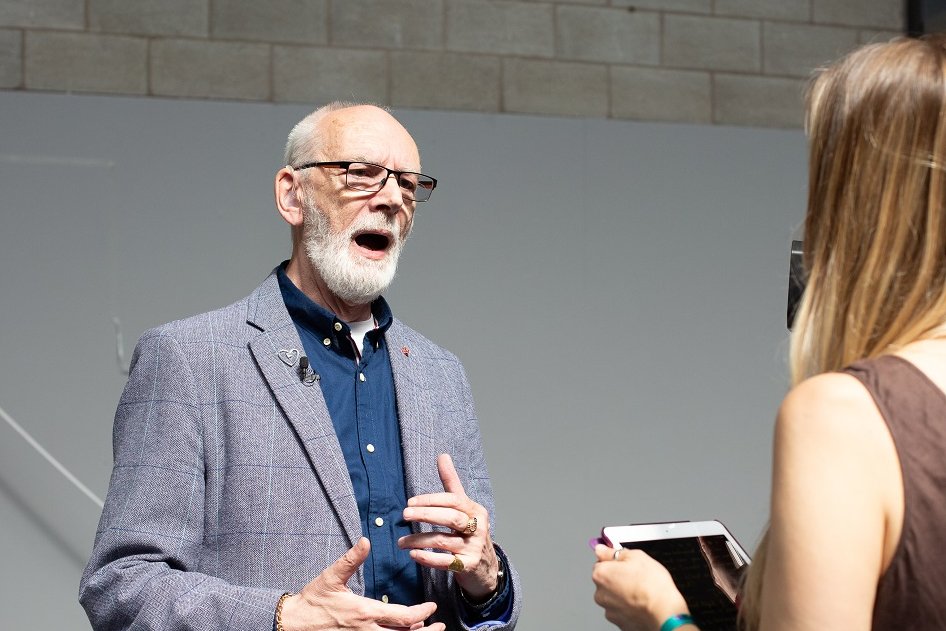 A white man with a beard and short white hair stands talking to a person interviewing him. He wears a blue shirt and jacket, and has a silver Terrence Higgins Trust heart pin logo. The interviewer has her back to the camera, she has long blonde hair and wears a brown linen dress.