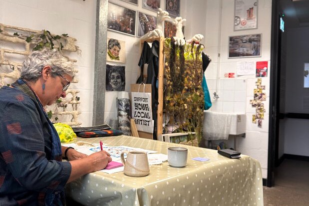 A white woman with grey hair sits drawing at a table in a studio with art on the walls. 