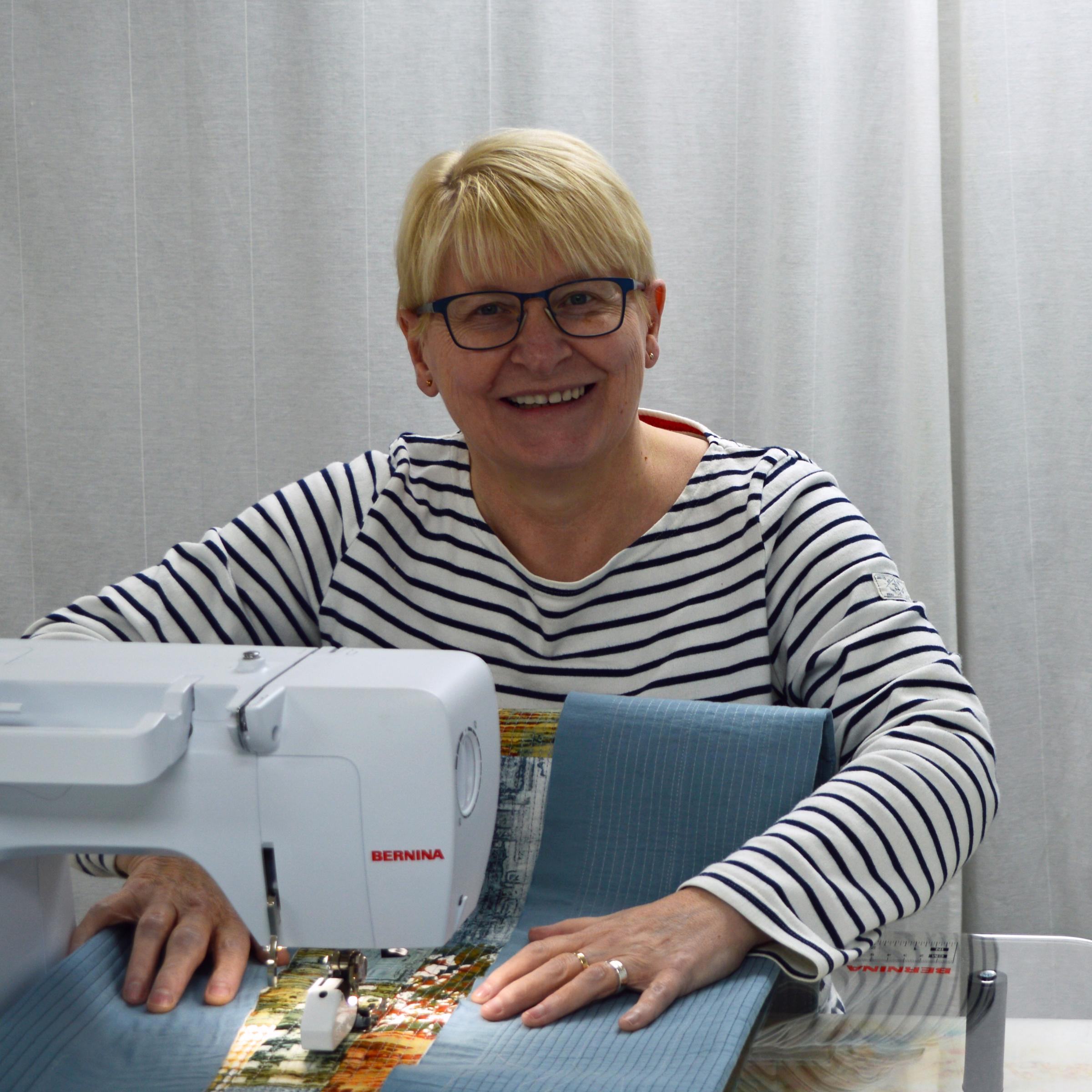 Image shows a white woman with short blonde hair sitting at her sewing machine which quilting a piece.