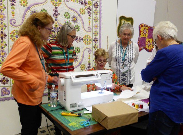 Woman sat an workbench with a sewing machine demonstrating to a group of women surrounding the table