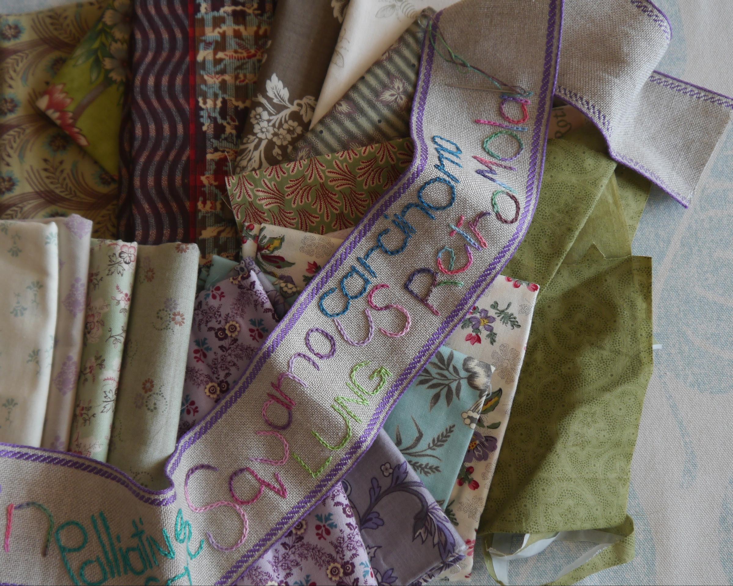 A bundle of green and purple floral patterned fabrics underneath a long narrow piece of beige fabric with purple edges with embroidered words on