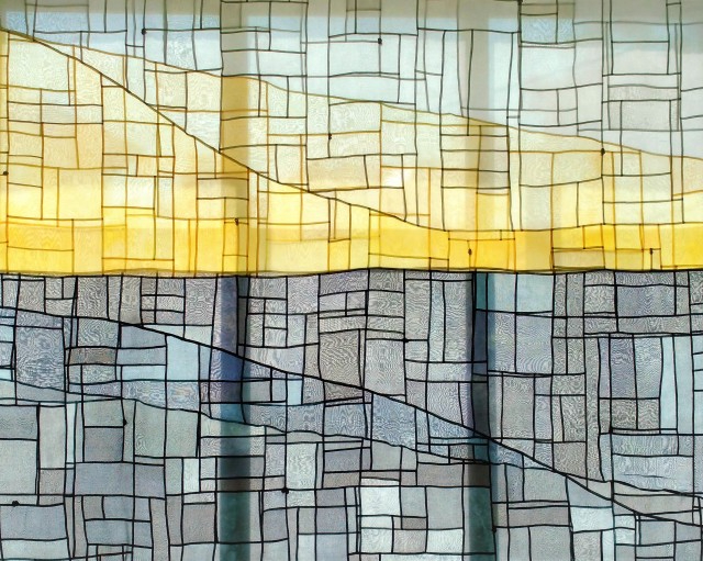 The Greyline, 2017 by Sara Cook. Hanging across a window with light coming through. Made of a light fabric in yellow and dark and light greys