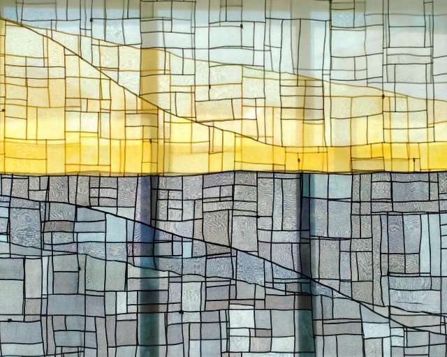 The Greyline, 2017 by Sara Cook. Hanging across a window with light coming through. Made of a light fabric in yellow and dark and light greys