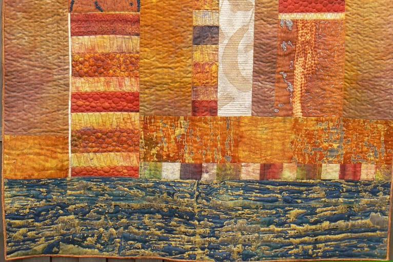 Close up of quilt made of textured fabrics with blue horizontal stripe at the bottom, and orange bronze, brown and beige rectangles above