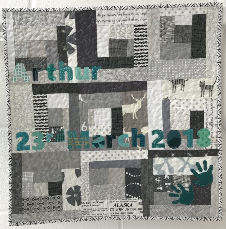 Modern style baby quilt made of grey and white rectangular patches with letters and numbers spelling 'Arthur' and '23rd March 2018' in green fabric on top. 