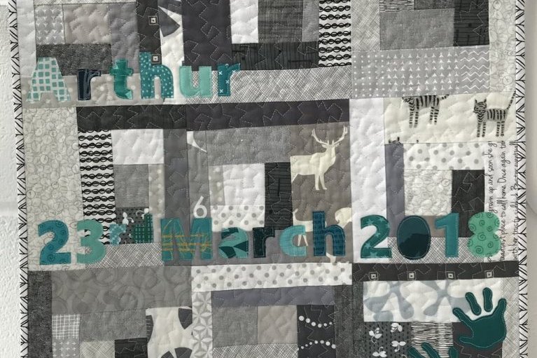 Modern style baby quilt made of grey and white rectangular patches with letters and numbers spelling 'Arthur' and '23rd March 2018' in green fabric on top. 