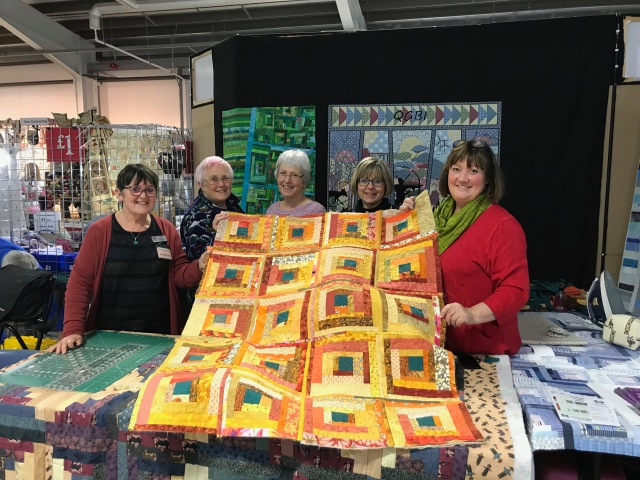 Hilary Jackson stood with a group of ladies holding up a yellow, orange and green quilt