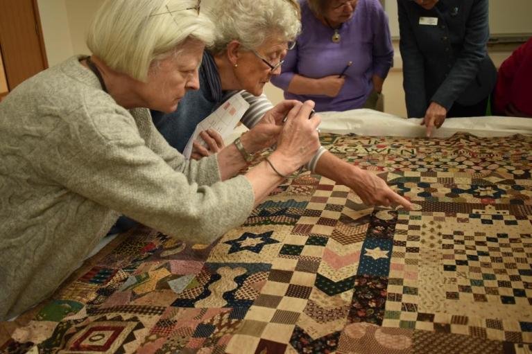Two ladies studying a quilted bedcover laid out on a table. One pointing at the quilt, the other taking a photo of it