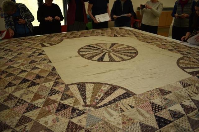 Patchwork quilt laid out on a table with a group of people stood around studying it.