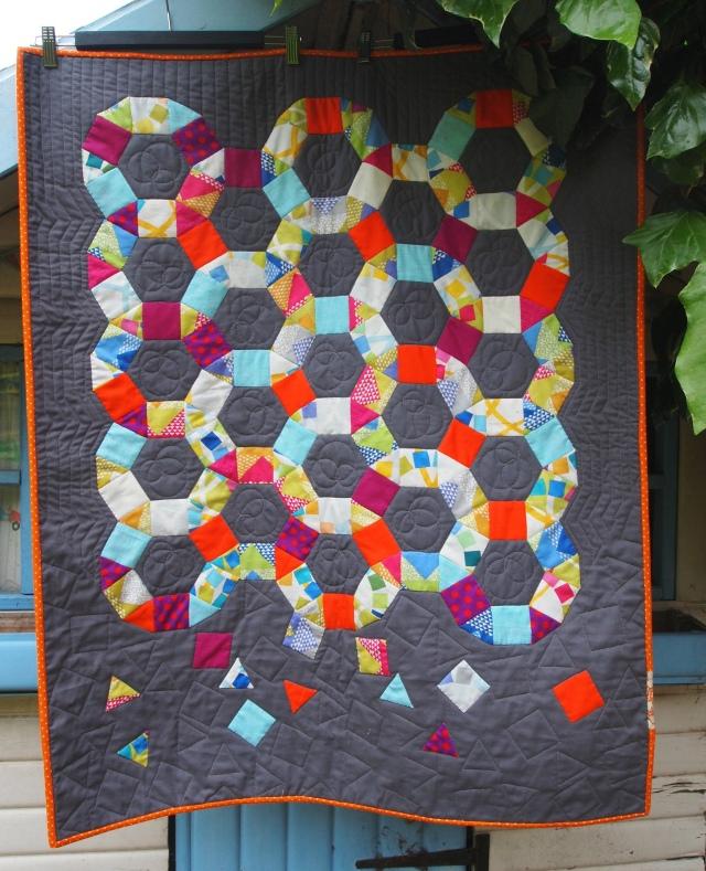 Large quilt with grey background and orange border, in the centre a design of interlocking rings made of multi-coloured fabric squares and triangles